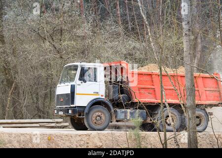 GRODNO, BÉLARUS - AVRIL 2020 : camion lourd de plusieurs tonnes MAZ avec cabine blanche et carrosserie orange, le long de la route de campagne poussiéreuse avec forêt de printemps Banque D'Images