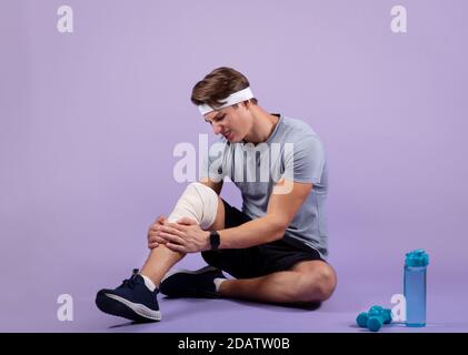 Portrait complet d'un jeune sportif ayant une blessure au genou, tenant sa jambe tendue en bandage, fond de studio violet Banque D'Images