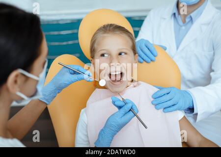 Gai enfant regarde le médecin assis dans la chaise, femme dentiste dans le masque de protection et les gants en caoutchouc commencent à traiter les dents Banque D'Images