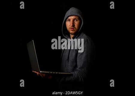 Un homme dans un capot tient un ordinateur portable dans ses mains sur un fond sombre. Concept de cyber-voyou, hacker. Bannière. Banque D'Images