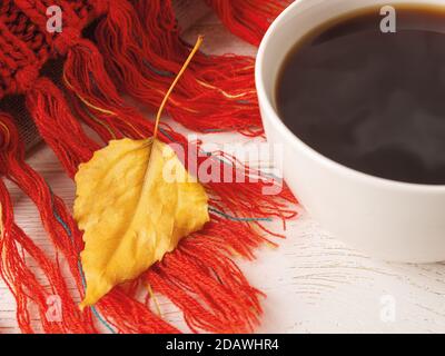 Gros plan sur une feuille d'automne jaune sèche sur une écharpe tricotée à franges rouges près d'une tasse blanche de café chaud à la vapeur. Ambiance automnale confortable. Mise au point sélective. Banque D'Images