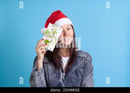 Portrait en gros plan d'une jeune femme asiatique heureuse dans une couronne de Noël qui cache un œil derrière une boîte cadeau, sur un fond bleu Banque D'Images