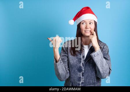 Maquette de la jeune femme asiatique découragée et péneuse surprise Chapeau de père Noël pointe à l'espace vide isolé sur fond bleu Banque D'Images