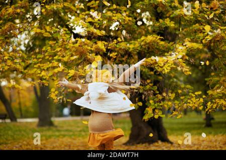 Bonjour automne. Vu de derrière une fille en chandail brun et chapeau orange se réjouissant dehors dans le parc de la ville en automne. Banque D'Images