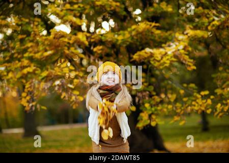 Bonjour automne. Jeune fille moderne souriante en chandail brun et chapeau orange jette des feuilles d'automne dehors dans le parc de la ville en automne. Banque D'Images