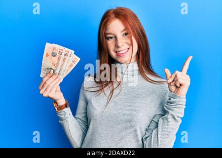 Jeune fille à tête rouge détenant 10 billets de banque au royaume-uni sourire heureux pointant avec la main et le doigt sur le côté Banque D'Images