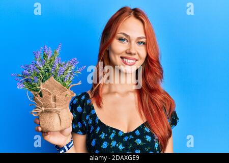 Jeune belle femme à tête rouge tenant pot de lavande regardant positif et se tenir debout et sourire avec un sourire confiant montrant les dents Banque D'Images