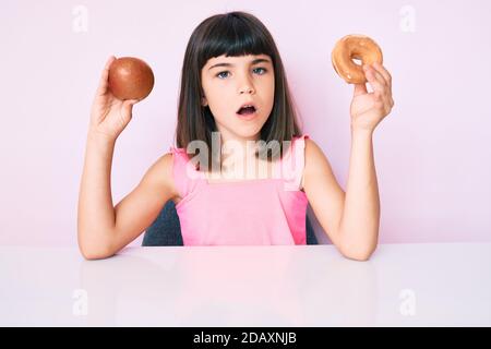 Jeune fille avec bang tenant pomme rouge et donut assis sur la table dans le visage de choc, regardant sceptique et sarcastique, surpris par la bouche ouverte Banque D'Images