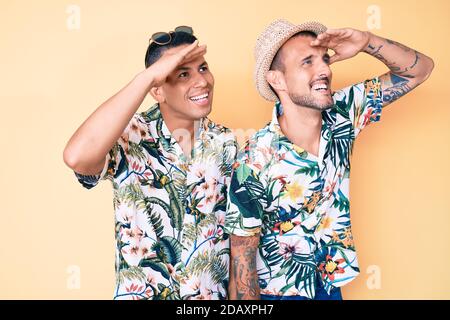 Jeune gay couple de deux hommes portant un chapeau d'été et une chemise hawaïenne très heureux et souriant regardant loin avec la main au-dessus de la tête. Concept de recherche. Banque D'Images