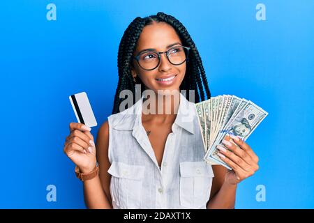 Belle femme hispanique tenant des dollars et carte de crédit souriant regardant le côté et regardant loin de penser. Banque D'Images