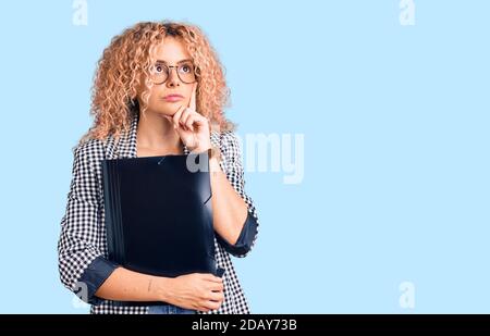 Jeune femme blonde avec des cheveux bouclés tenant un dossier d'affaires sérieux visage pensant à la question avec la main sur le menton, réfléchi à l'idée de confusion Banque D'Images