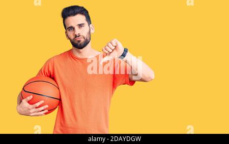 Jeune homme beau avec la barbe hailding ballon de basket-ball avec visage en colère, signe négatif montrant l'aversion avec les pouces vers le bas, concept de rejet Banque D'Images
