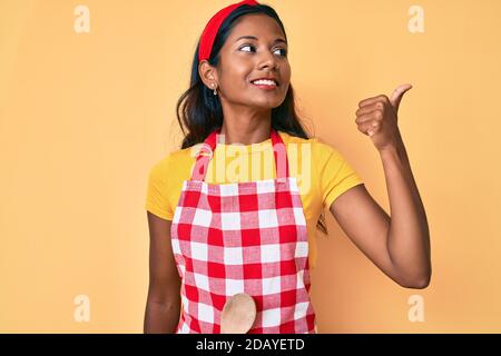 Jeune fille indienne portant un tablier de boulanger pointant le pouce vers le haut le côté souriant heureux avec la bouche ouverte Banque D'Images