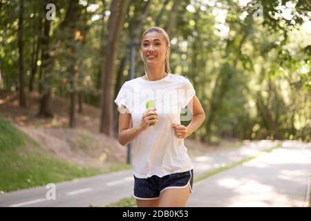 Femme écouter de la musique courir sur la piste d'asphalte parc d'été Caucasien Ecouteurs femelles jogging matin ensoleillé entraînement de coureur tenir le smartphone dans Talon de la main Banque D'Images