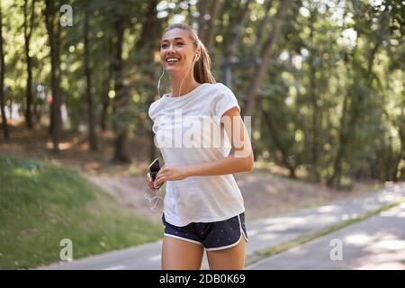 Femme écouter de la musique courir sur la piste d'asphalte parc d'été Caucasien Ecouteurs femelles jogging matin ensoleillé entraînement de coureur tenir le smartphone dans Talon de la main Banque D'Images