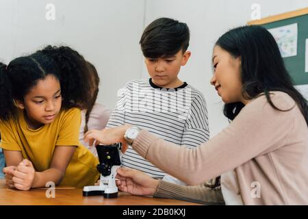 Jeune femme montrant des enfants multiethniques microscope en cours de science, d'apprentissage, de découverte, de curiosité. Professeur d'école asiatique aidant les élèves à utiliser le microscope Banque D'Images