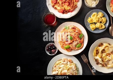Cuisine italienne, plan de travail à plat. Pâtes, olives, vin sur fond noir avec espace de copie. Spaghetti à la sauce tomate, raviolis et autres Banque D'Images