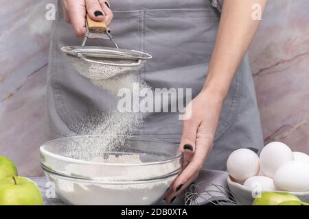 Femme tamisant la farine à travers le tamis, préparant une tarte douce maison avec des pommes mûres Banque D'Images