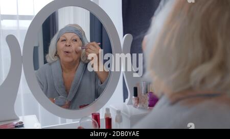 Vieille femme grand-mère prenant soin de la peau, appliquant la poudre de fondation anti-rides avec le pinceau, mettant le maquillage sur. Grand-mère âgé effectuant des procédures de maquillage. Cosmétiques pour retraités Banque D'Images