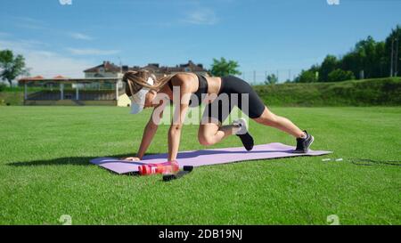 Jeune femme musclée portant un chapeau blanc faisant de l'exercice alpiniste sur le tapis en été ensoleillé. Magnifique entraînement de fille muscles de base sur le terrain de stade sur l'herbe verte fraîche. Concept d'entraînement. Banque D'Images