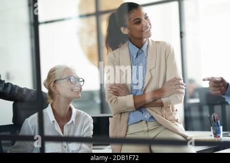 Deux femmes souriantes différentes collègues regardant de côté tout en ayant un réunion avec des collègues dans le bureau moderne Banque D'Images