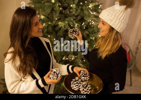 Deux belles femmes souriantes se regardant l'une l'autre tout en mettant pommes de pin blanches sur un arbre de noël Banque D'Images