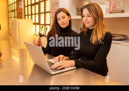 Deux belles femmes portant un col de cygne noir, travaillant avec un ordinateur portable dans la cuisine et prenant une tasse de vin rouge Banque D'Images