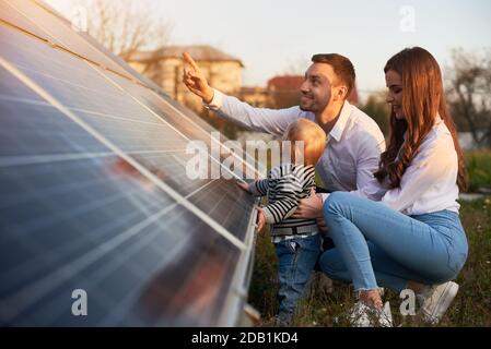 Vue latérale d'une jeune famille moderne avec un petit garçon se familiarisant avec le panneau solaire lors d'une journée ensoleillée, concept d'énergie alternative verte Banque D'Images