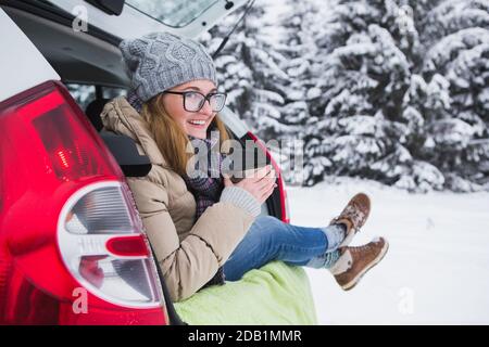 La femme est assise dans le coffre de la voiture et tient une tasse de thé chaud dans ses mains. Banque D'Images