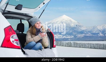 La femme est assise dans le coffre de la voiture et tient une tasse de thé chaud dans ses mains. Banque D'Images