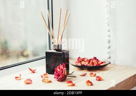 Récipient en verre brun avec diffuseur en bâton de bois sur le seuil de fenêtre en bois de la maison avec de beaux pétales de rose secs pour la décoration. Banque D'Images