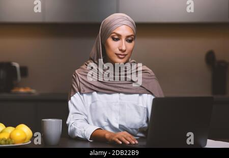 Femme musulmane moderne travaillant sur ordinateur portable en ligne assis dans la cuisine Banque D'Images