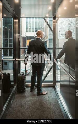 Homme d'affaires avec valise dans le centre d'affaires. Concept de voyage d'affaires. Un homme de race blanche d'âge moyen regarde par la paroi en verre de l'ascenseur. Vue arrière Banque D'Images
