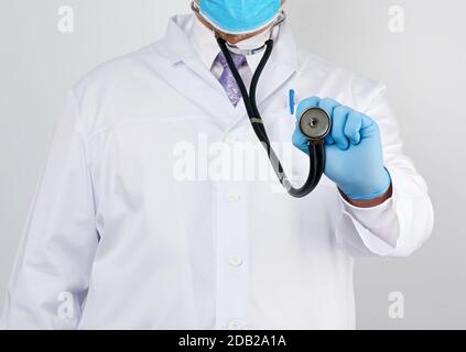 médecin dans un manteau blanc et attache dans sa main un phonendoscope noir, portant des gants stériles bleus, fond blanc Banque D'Images