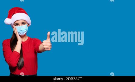 Femme excitée et choquée portant chapeau de père Noël et masque médical de protection du visage montrant le pouce vers le haut geste, arrière-plan bleu isolé. Banque D'Images