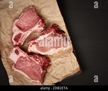 tranches de porc juteux cru de viande sur la côte, la nourriture se trouve sur du papier parchemin brun, fond en bois noir, vue de dessus Banque D'Images