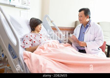 Médecin asiatique confiant explantant à une patiente gaie sur un état de santé allongé sur le lit à l'hôpital. Banque D'Images