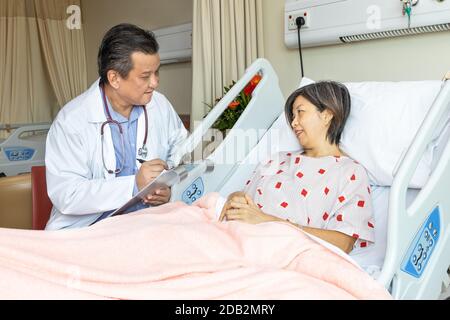 Médecin asiatique confiant explantant à une patiente gaie sur un état de santé allongé sur le lit à l'hôpital. Banque D'Images