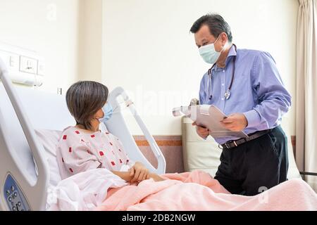 Médecin asiatique confiant avec masque facial explantant à une patiente gaie sur un état médical allongé sur le lit à l'hôpital. Banque D'Images