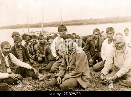 Hommes et garçons indiens vêques de l'Ouest, assis le long du rivage, bateaux en arrière-plan. Banque D'Images