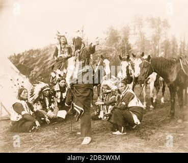 Les Indiens du Dakota, peut-être membres du spectacle Wild West de Pawnee Bill, assis en demi-cercle, avec des chevaux et des tipi, un homme à cheval derrière eux, et un autre homme debout et pointant. Banque D'Images