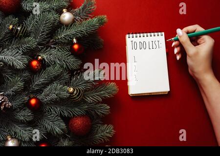 Une femme écrit une liste de shopping ou de tâches dans un carnet sur un fond rouge à côté des branches et des décorations du nouvel an. Maquette. Banque D'Images