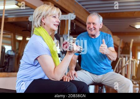 Femme âgée dans la salle de gym s'exerçant avec des haltères pour la forme physique tandis que son mari la met au feu Banque D'Images