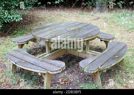 Table ronde de pique-nique en bois avec sièges arrondis sur l'herbe avec sol en dessous et arbustes en arrière-plan. Banque D'Images