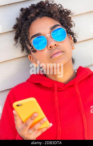 Fille adolescente cool adolescent mixte race biracial Afro-américaine femelle jeune femme portant des lunettes de soleil bleues et un sweat à capuche rouge utilisant téléphone portable en vacances Banque D'Images