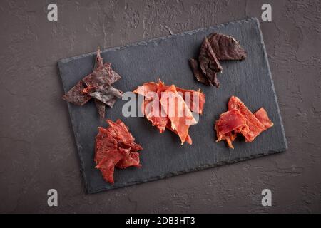 Saccadé. Ensemble de divers types de viande épicée séchée sur une planche à découper en pierre noire sur fond gris foncé. Vue de dessus. En-cas pour la bière. Banque D'Images