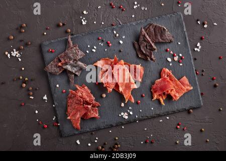 Saccadé. Ensemble de divers types de viande épicée séchée sur une planche à découper en pierre noire sur fond gris foncé. Vue de dessus. En-cas pour la bière. Banque D'Images