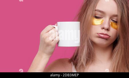 Femme fatiguée et ivre avec la gueule de bois et les maux de tête mettre des taches sur les yeux tenant une tasse de café blanc, isoler le fond rose Banque D'Images