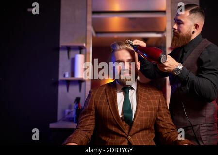 homme élégant assis dans le salon de coiffure pendant que coiffeur sèche ses cheveux, beau jeune homme caucasien barbu heureux obtenant une coupe de cheveux à la mode, barber servant le client Banque D'Images