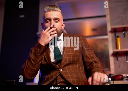 homme caucasien riche et confiant avec barbe en costume élégant, asseyez-vous fumer du cigare, détendez-vous. isolé dans une pièce fumée sombre Banque D'Images